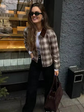 Woman in plaid shirt and black pants standing against a gray wall.